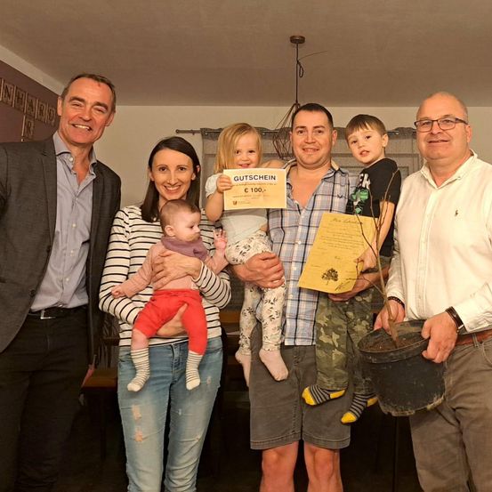 Fünf Erwachsene und drei Kinder stehen in einem Raum und lächeln. Ein Mann hält ein Zertifikat, und ein anderer hält einen Topf mit einer Pflanze. Sie scheinen einen besonderen Anlass zu feiern.