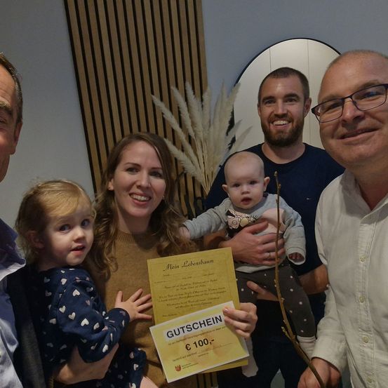 Eine Familie mit fünf Personen posiert für ein Foto. Die Frau hält ein Zertifikat und ein Baby. Der Mann rechts trägt eine Brille und lächelt. Der Mann in der Mitte hält ein Baby mit einem Band. Das Baby schaut in die Kamera.