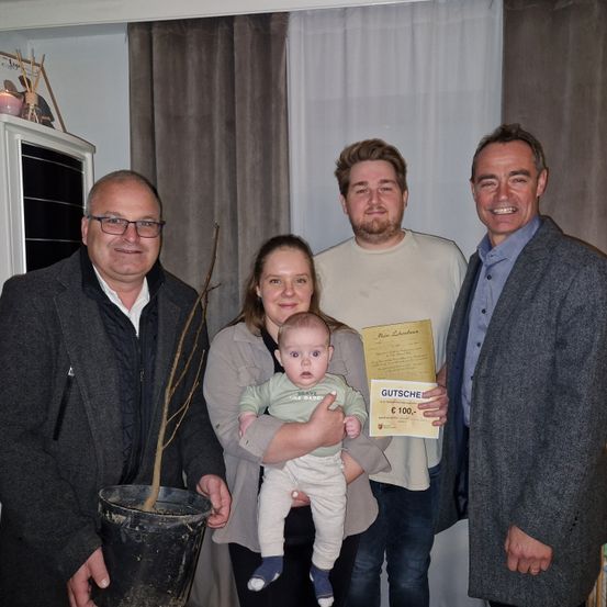 Fünf Menschen stehen lächelnd für ein Foto im Innenraum. Ein Baby in einem grünen Shirt wird von einer Frau in einer grauen Jacke gehalten. Ein Mann auf der linken Seite hält eine Topfpflanze. Ein anderer Mann auf der rechten Seite hält ein Zertifikat.