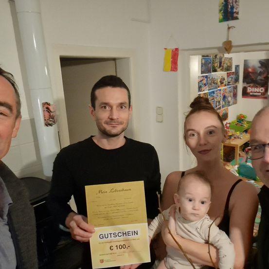 Vier Personen stehen in einem Raum und lächeln für ein Foto. Ein Mann hält ein Zertifikat und ein Baby ist in den Armen einer Frau. Ein anderer Mann ist links und eine Flagge hängt an der Wand.