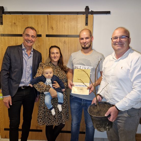 Vier Personen stehen zusammen in einem Raum. Der Mann auf der linken Seite hält ein Baby. Der Mann in der Mitte hält eine Topfpflanze und ein Zertifikat. Die Frau in der Mitte hält das Baby. Der Mann auf der rechten Seite trägt eine Brille.