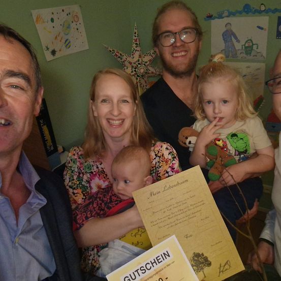 Eine Familie posiert für ein Foto mit zwei Babys. Ein Baby wird von einer Frau gehalten und das andere wird von einem Mädchen getragen. Sie alle lächeln und schauen in die Kamera. Hinter ihnen ist eine grüne Wand mit einigen Zeichnungen und einer Sternendekoration.