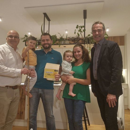 Fünf Menschen posieren lächelnd für ein Foto in einer Küche mit Holzboden. Ein Mann hält ein gelbes Zertifikat und ein anderer ein Baby.