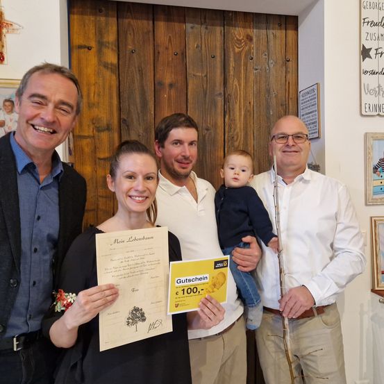 Eine vierköpfige Familie posiert für ein Foto. Eine Frau hält ein Zertifikat, ein Mann ein Baby, und zwei Männer stehen neben ihnen. Der Mann rechts hält eine Flöte. Sie sind drinnen mit Holzwänden und Bilderrahmen.