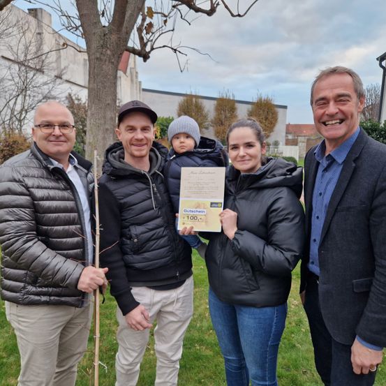 Eine Familie und zwei Männer stehen in einem Park. Sie alle lächeln und scheinen für ein Foto zu posieren. Der Mann auf der linken Seite hält einen Stock. Die Familie in der Mitte hält ein Zertifikat. Hinter ihnen steht ein Baum und ein Gebäude.