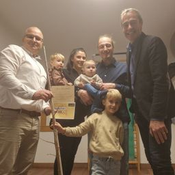 Eine Familie mit sechs Personen, darunter zwei kleine Kinder, steht gemeinsam in einem Raum mit Holzboden und weißen Wänden. Ein Mann hält ein Zertifikat und einen Stock, während ein anderer Mann ein Baby hält.