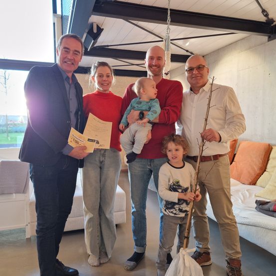 Fünf Menschen posieren drinnen für ein Foto. Ein Mann hält ein Papier und ein Baby, während ein anderer Mann einen Stock hält. Ein Mann im Anzug lächelt und ein kleiner Junge hält einen Papierbeutel.