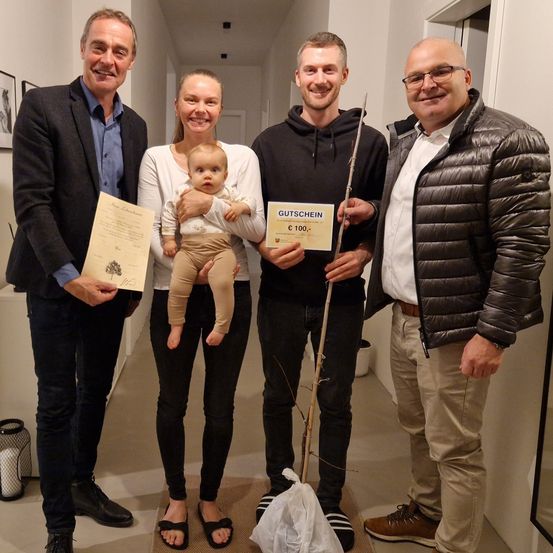 Vier Erwachsene stehen in einem Flur, zwei Männer und zwei Frauen. Eine Frau hält ein Baby, während die andere ein Zertifikat in der Hand hat. Ein Mann hält ein Zertifikat und einen Stock, der andere ein Zertifikat. Sie alle lächeln.