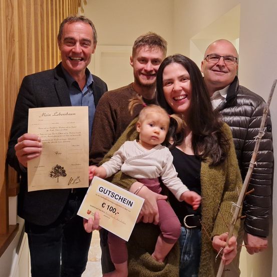 Eine fünfköpfige Familie posiert drinnen für ein Foto. Ein Mann hält ein Zertifikat und einen Gutschein. Die Frau hält ein Baby und lächelt. Dahinter steht ein Mann und hält einen Stock.
