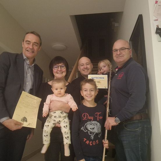 Eine Familie mit fünf Mitgliedern, darunter zwei Kinder, posiert für ein Foto in einem Flur. Der Mann auf der linken Seite hält ein Zertifikat, während die anderen ein Zertifikat und ein Schild halten.