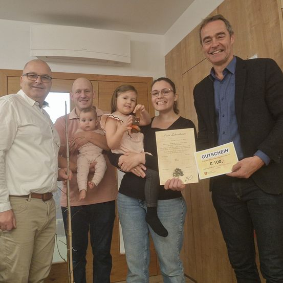 Fünf Personen stehen in einem Raum, lächeln und posieren für ein Foto. Zwei Erwachsene halten ein Baby und ein Kind, und ein Mann hält ein Papier und ein Zertifikat.