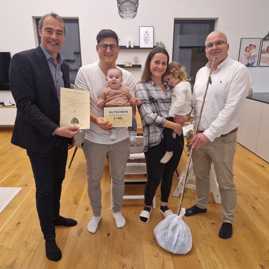 Vier Personen stehen in einem Raum zusammen. Zwei Männer halten ein Zertifikat und eine Geschenkkarte. Eine Frau hält ein Baby und ein Kleinkind. Ein anderer Mann hält einen Stock mit einem Beutel.