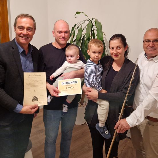 Fünf Erwachsene und zwei Kinder posieren für ein Foto in Innenräumen. Der Mann auf der linken Seite hält ein Zertifikat. Der Mann in der Mitte hält ein Baby, und die Frau neben ihm hält ein weiteres Baby. Der Mann auf der rechten Seite hält einen Stock. Hinter ihnen steht eine Topfpflanze.