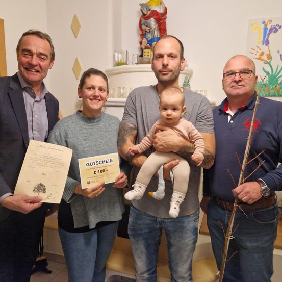 Eine Familie und zwei Männer stehen in einem Raum und halten ein Zertifikat und einen Gutschein. Der Mann auf der linken Seite hält ein Zertifikat, der Mann auf der rechten Seite einen Gutschein. Der Mann in der Mitte hält ein Baby. Hinter ihnen befinden sich Gemälde und ein Regal.