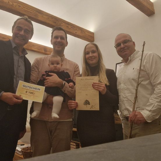 Vier Personen, zwei Männer und zwei Frauen, stehen in einem Raum. Sie halten ein Zertifikat und einen Gutschein. Der Mann auf der linken Seite hält einen Gutschein im Wert von 100 Euro. Die Frau auf der rechten Seite hält ein Zertifikat mit einer Baumzeichnung. Das Baby wird vom Mann in der Mitte gehalten.