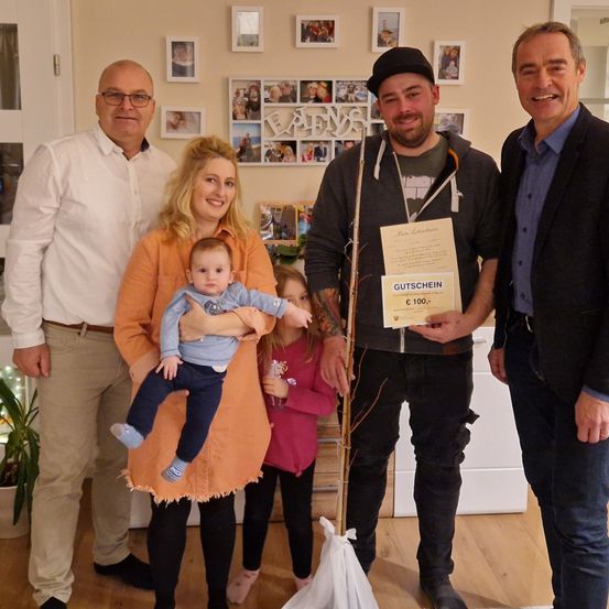 Fünf Menschen posieren für ein Foto in einem Raum, halten eine Pflanze, ein Zertifikat und eine Handtasche. Der Mann auf der rechten Seite lächelt und der Mann auf der linken Seite schaut in die Kamera. Hinter ihnen hängen Bilderrahmen an der Wand.