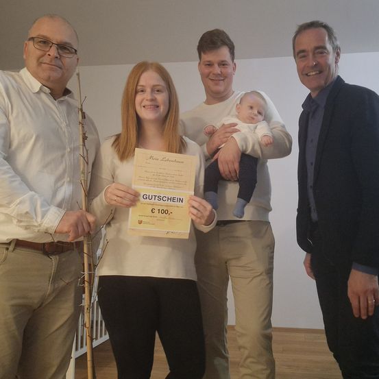 Vier Menschen stehen lächelnd in einem Raum. Eine Frau hält ein Zertifikat und ein Mann ein Baby. Hinter ihnen befindet sich eine weiße Wand und ein Holzboden.