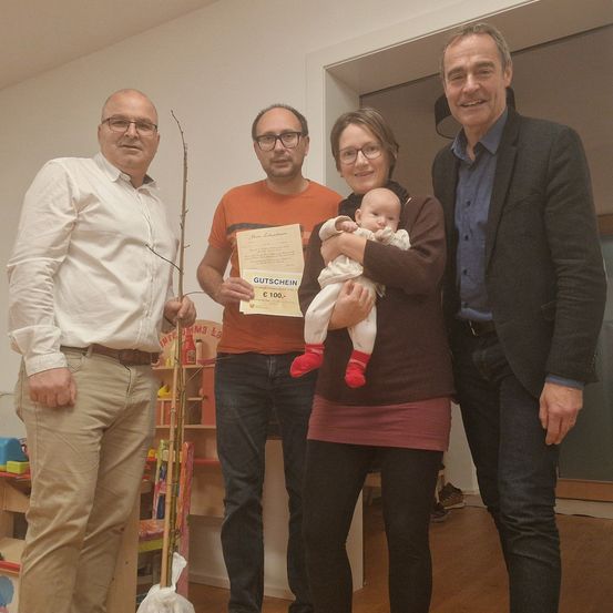 Vier Personen stehen in einem Raum, eine Frau hält ein Baby. Ein Mann links hält einen Stock, und ein anderer Mann hält ein Zertifikat. Dahinter befindet sich ein Regal mit Spielzeug.