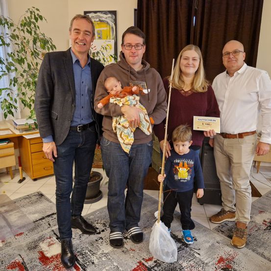 Eine fünfköpfige Familie posiert für ein Foto in einem Raum mit Teppich, Topfpflanze und Möbeln. Der Mann in der Mitte hält ein Baby, während die Frau eine Angelrute hält. Der Mann auf der rechten Seite hält ein Zertifikat.