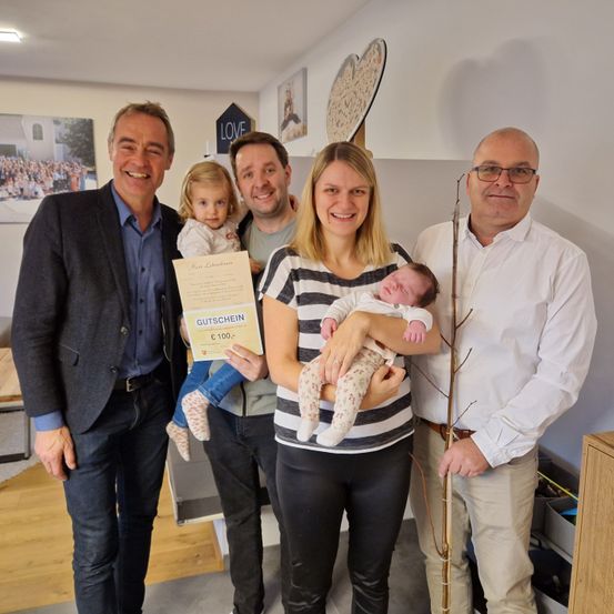 Eine Familie mit vier Personen posiert für ein Foto in einem Raum mit einem Baum, einem Geschenkgutschein und einem dekorativen Herz an der Wand. Ein Mann und eine Frau halten ein Baby, und ein anderer Mann hält ein kleines Mädchen.