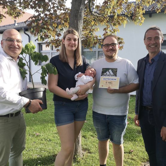 Fünf Personen stehen lächelnd in einem Garten. Eine Frau hält ein Baby und ein Mann einen Topf mit einer Pflanze. Ein anderer Mann hält ein Zertifikat. Hinter ihnen steht ein Haus mit Glasfenstern.