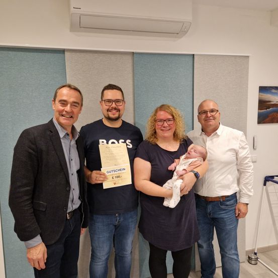 Eine Gruppe von vier Personen steht eng beieinander im Innenbereich. Eine Frau hält ein Baby, während ein Mann ein Zertifikat in der Hand hat. Sie alle lächeln für die Kamera. Dahinter befindet sich eine Wand mit einer Klimaanlage und einem Gemälde.