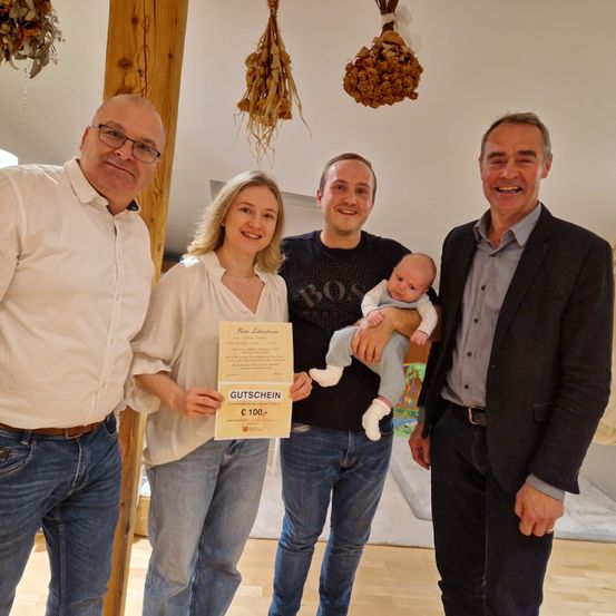 Eine Familie mit vier Personen und einem Baby posiert für ein Foto in einem Raum mit Holzboden und Dekorationen. Die Frau hält ein Zertifikat und der Mann das Baby.