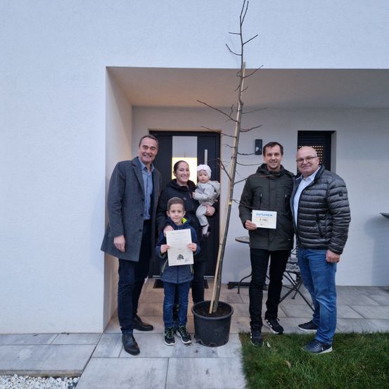 Eine Familie posiert für ein Foto vor einem Haus mit einem Topfbaum. Der Mann links hält ein Zertifikat. Das Baby wird von einer Frau in der Mitte gehalten. Ein anderer Mann hält ein Zertifikat auf der rechten Seite.