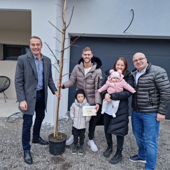 Fünf Personen stehen vor einem Gebäude und lächeln für ein Foto. Ein Mann hält einen jungen Baum und eine Frau ein Baby in einer rosa Mütze. Hinter ihnen hält ein weiterer Mann ein Zertifikat.