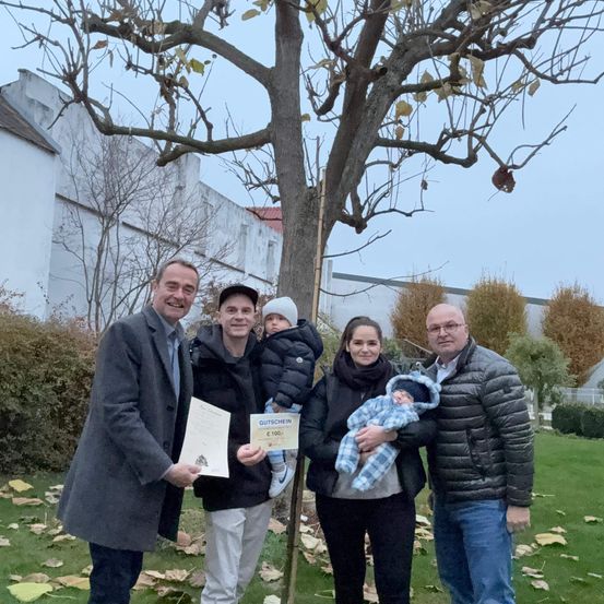 Eine Familie steht um einen Baum in einem Park, mit zwei Männern und zwei Frauen, eine hält ein Baby und eine andere ein Zertifikat.