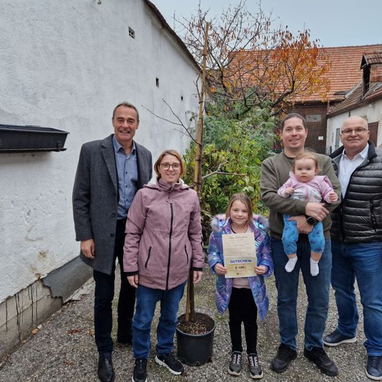 Eine Gruppe von Menschen steht zusammen draußen. Sie lächeln und posieren für ein Foto. Ein junges Mädchen hält ein Zertifikat. Hinter ihnen steht ein Topfbaum. Im Hintergrund befinden sich Gebäude.