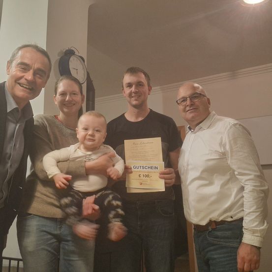 Vier Menschen stehen in einem Raum und lächeln. Eine Frau hält ein Baby, während ein Mann ein Zertifikat mit einem Geschenkgutschein für 100 Euro in der Hand hat. Dahinter befindet sich eine Uhr.
