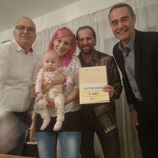 Fünf Personen posieren für ein Foto im Innenraum. Eine Frau hält ein Baby und einen Gutschein. Der Mann rechts hält einen Gutschein mit Logo.