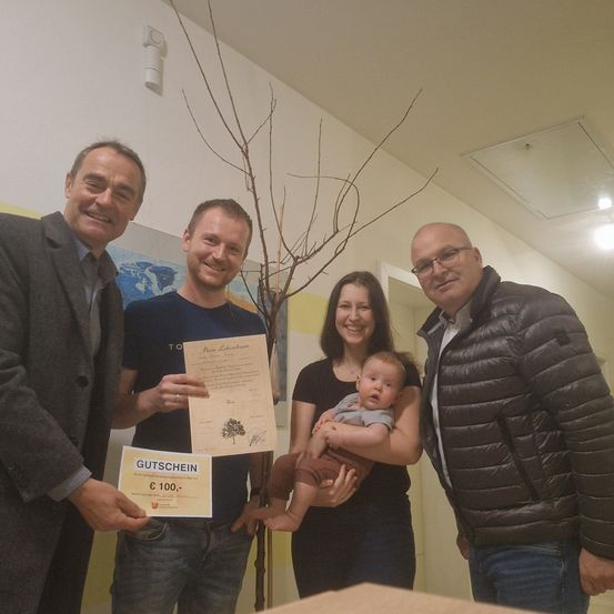 Vier Personen posieren für ein Foto. Ein Mann links hält ein Zertifikat, während eine Frau in der Mitte ein Baby im Arm hält. Ein Mann rechts trägt eine Brille und einen Mantel. Sie befinden sich in einem Raum mit weißer Wand und Decke.