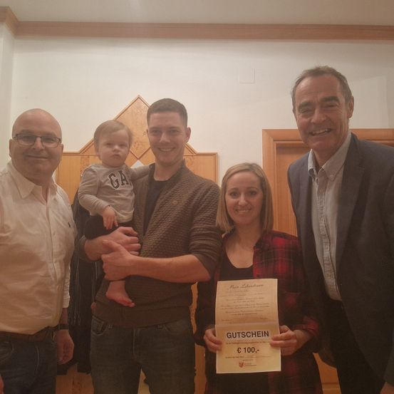 Fünf Personen stehen in einem Raum, halten ein Zertifikat und lächeln. Ein Baby wird von einem Mann gehalten. Eine Frau hält ein Zertifikat.