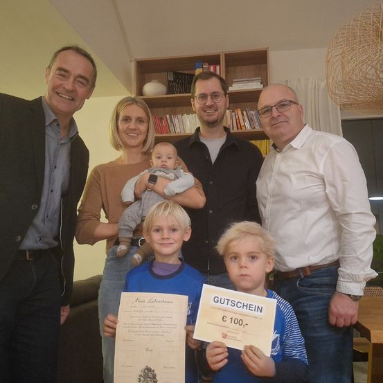 Eine Familie von fünf Personen steht zusammen in einem Raum mit Bücherregalen und einer Lampe. Sie lächeln für ein Foto. Ein Baby, ein Junge und ein Mädchen halten Urkunden. Der Junge hält eine Urkunde mit dem Text 'Gutschein' und 'C 100,-'.