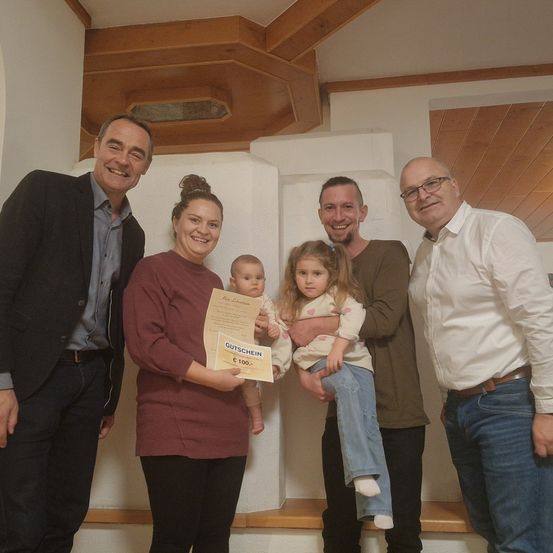 Eine vierköpfige Familie steht in einem Raum mit einer Holzdecke und Wänden. Die Frau hält ein Baby und einen Gutschein, während der Mann neben ihr ein weiteres Kind hält. Zwei andere Männer stehen auf beiden Seiten und lächeln.