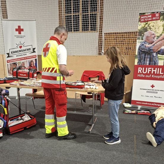 Ein Mann und eine Frau stehen an einem Tisch und bereiten sich möglicherweise auf eine Erste-Hilfe-Schulung vor. Der Mann trägt eine Warnweste, die Frau eine Kapuzenjacke. Verschiedene Ausrüstungsgegenstände liegen auf dem Tisch. Dahinter befindet sich ein Poster zur Ersten Hilfe.