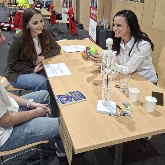Zwei Frauen und ein Mann sitzen an einem Tisch mit einem menschlichen Skelett und Büchern. Die Frau in Weiß hält ein Modell des menschlichen Herzens.