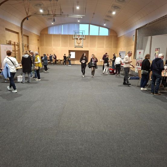 Ein großer Saal mit stehenden und gehenden Personen. Einige tragen Taschen. Es gibt aufgestellte Tische und Stühle. Ein Basketballkorb ist in der Ferne sichtbar.