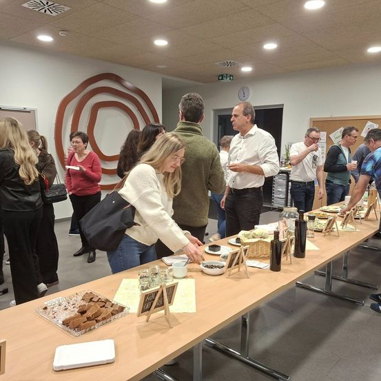 Eine Gruppe von Menschen versammelt sich in einem Raum mit einem langen Tisch voller Essen. Eine Frau in einer weißen Strickjacke serviert Essen. Ein Mann in einem weißen Hemd spricht mit einem anderen Mann.