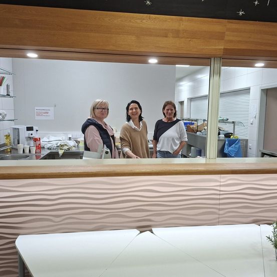 Drei Frauen stehen hinter einer Theke in einer Küche. Eine Frau trägt eine Brille. Ein Waschbecken und eine Kaffeemaschine stehen hinter ihnen. Eine kleine Pflanze steht auf dem Tisch.