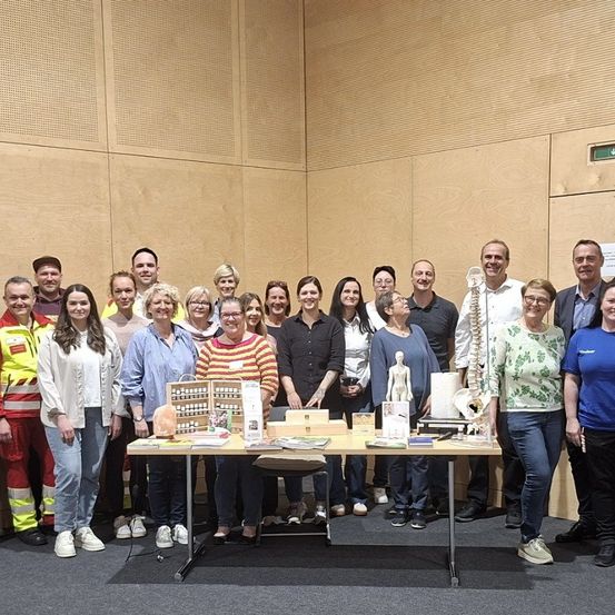 Gruppe von Menschen, die um einen Tisch mit verschiedenen Gegenständen, einschließlich eines menschlichen Skeletts und einer Figur, in einem Raum mit einer beigefarbenen Wand stehen.