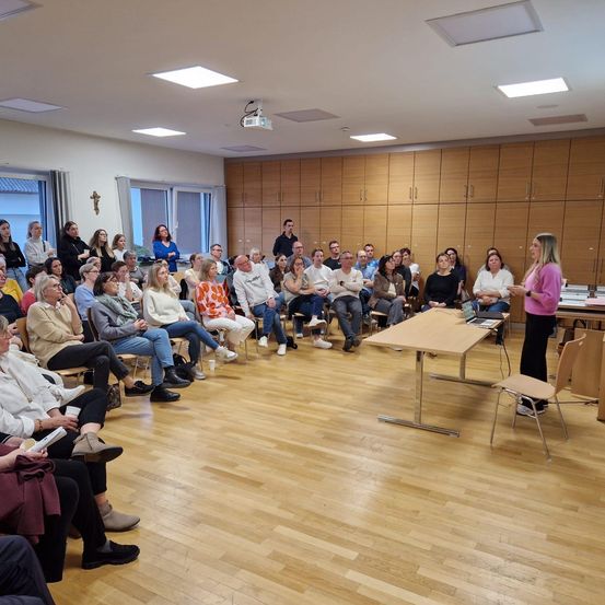 Eine Gruppe von Menschen sitzt in Stühlen vor einer Frau, die vor einem Tisch mit Laptop und Beamer steht. Der Raum hat Holzböden und Schränke.