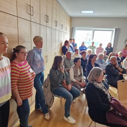 Eine Gruppe von Erwachsenen sitzt im Kreis in einem Raum mit Holzböden. Mehrere Frauen stehen in der Nähe einer Wand mit Holzschränken. Der Raum hat ein großes Fenster und eine Decke mit Lichtern.