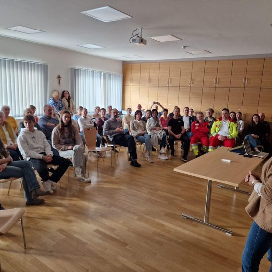 Ein Raum mit Holzböden und weißen Wänden. Menschen sitzen auf Stühlen, eine Frau steht vorne. Ein Schreibtisch mit Laptop und eine Tafel. Holzschränke an der Wand.