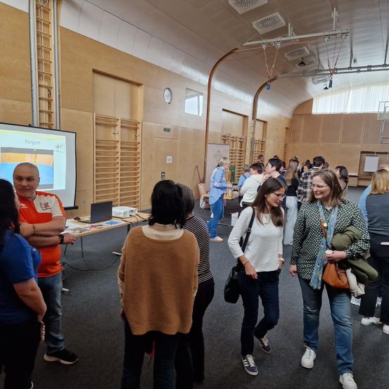 Menschen versammeln sich in einem Konferenzraum mit einer Projektionsleinwand, einige unterhalten sich. Ein Mann in Orange steht bei einem Tisch mit einem Laptop.