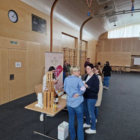 Eine Gruppe von Menschen versammelt sich um einen Tisch in einem Saal und betrachtet Weinflaschen. Ein Schild an der Wand steht für 'Bio Schäfer'. Eine Uhr und ein Whiteboard sind ebenfalls sichtbar.