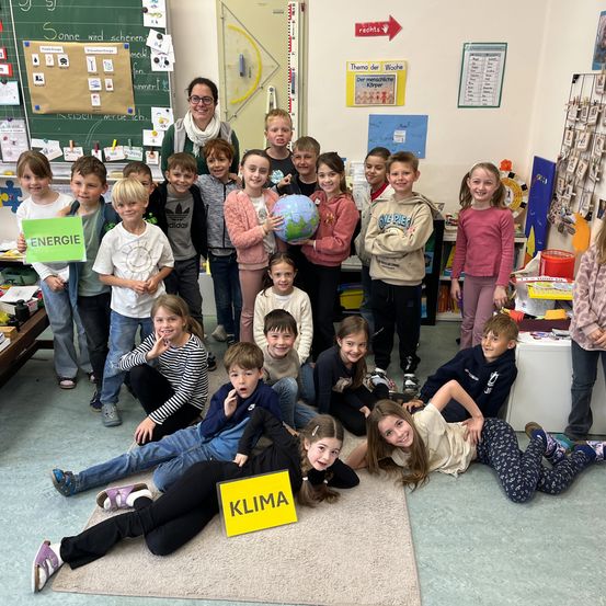 Ein Klassenzimmer voller Kinder mit Schildern Energie und Klima, einer Lehrerin und einem Globus in der Mitte.