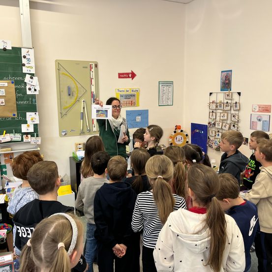 Eine Frau, die ein Buch hält, steht vor einer Klasse von Kindern. Der Klassenraum ist mit verschiedenen Postern und Diagrammen an den Wänden geschmückt.
